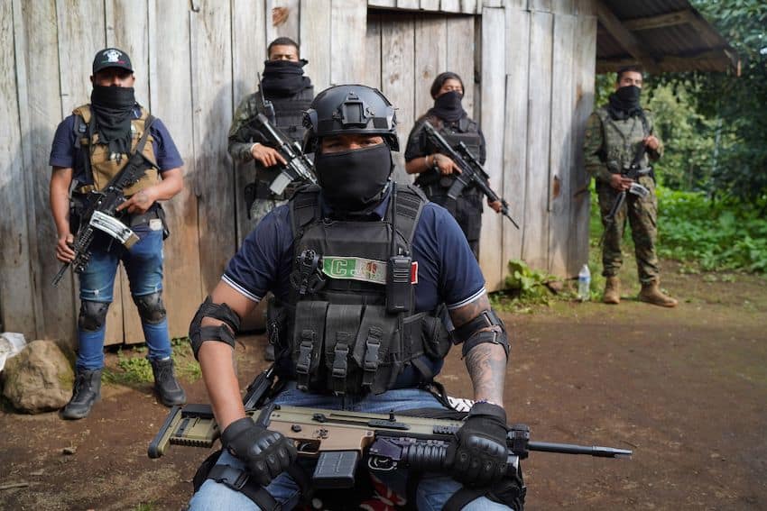 A man with his face covered wearing military gear labeled "CJNG" and carrying an assault rifle, with similarly dressed civilians standing behind him.