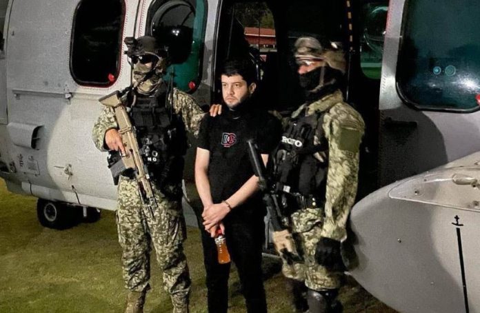 El Nini, a man in his 30s or 40s, stands between two Mexican soldiers in front of a helicopter.