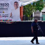 A federal agent patrols a plaza with a banner for murdered mayoral candidate José Alfredo Cabrera Barrientos in the background.