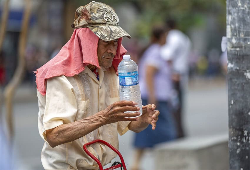 A street vendor wearing a hat and shade cloth over his neck offers a bottle of water.