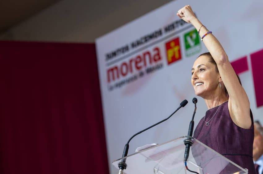 Claudia Sheinbaum stands at a podium in front of a sign showing the logos of Morena and its allies