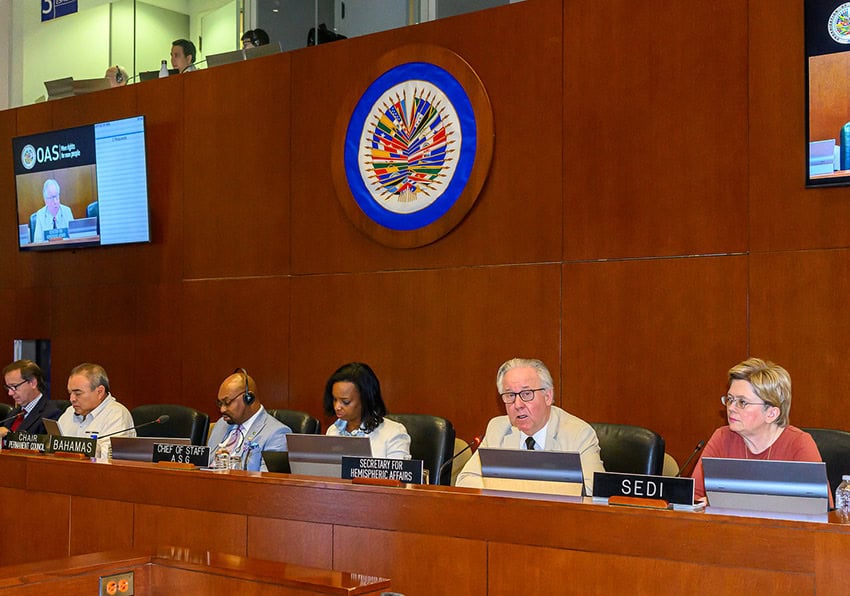 Representatives with placards reading SEDI, Secretary for Hemispheric Affairs, Chair Permanent Council and more sit at a wooden conference stage with microphones and the OAS logo.